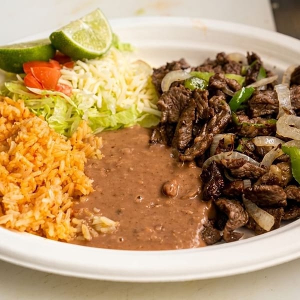 A plate of carne asada with rice, refried beans, lettuce, cheese, tomatoes, and lime wedges.