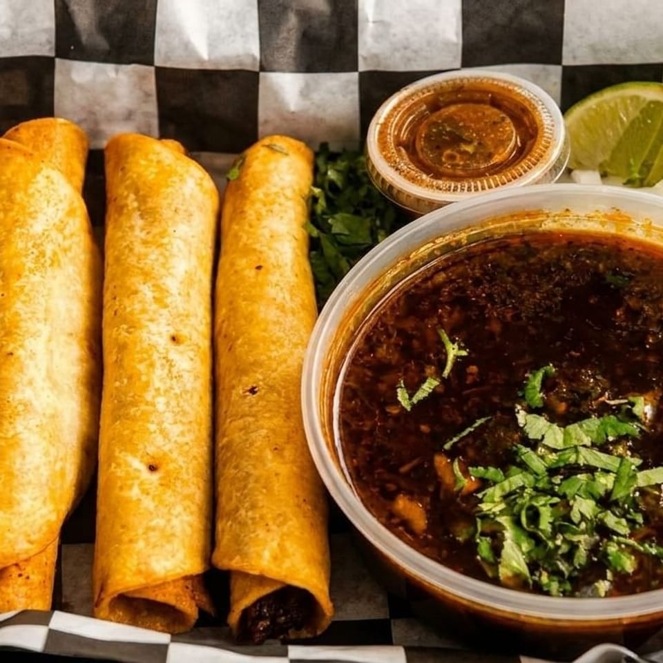 Four golden taquitos with a side of rich birria broth, lime, and salsa on checkered paper.